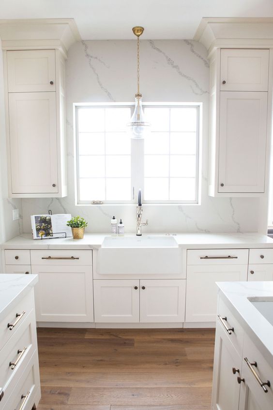an airy white kitchen with shaker cabinets, a white quartz backsplash and countertops plus brass fixtures is wow