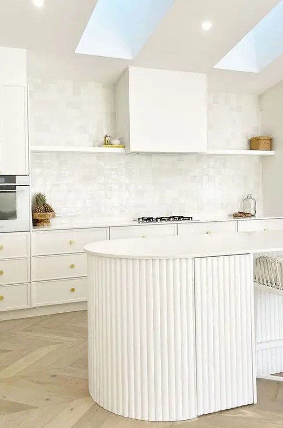 an airy white kitchen with white cabinets and an open shelf, a curved fluted kitchen island and skylights over it