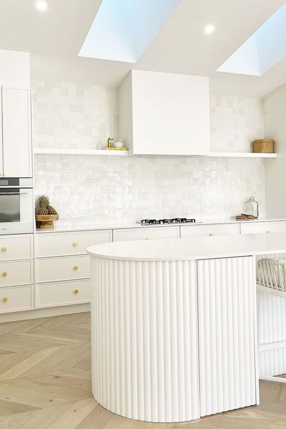 an airy white kitchen with white cabinets and an open shelf, a curved fluted kitchen island and skylights over it