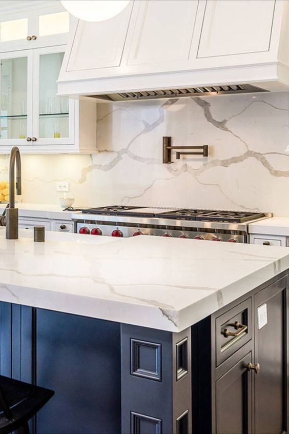 an elegant kitchen with white cabinets and a navy kitchen island, a white quartz backsplash and countertops plus a white hood