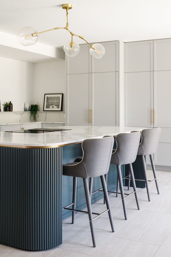 an exquisite dove grey kitchen with shaker cabinets, white marble countertops and a backsplash, a navy fluted kitchen island with a cooking and sitting zone