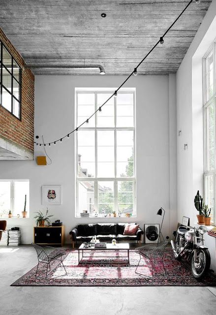 a boho double-height living room with a wooden ceiling, double-height windows, a black sofa, a coffee table, metal chairs, a bright rug and a bike