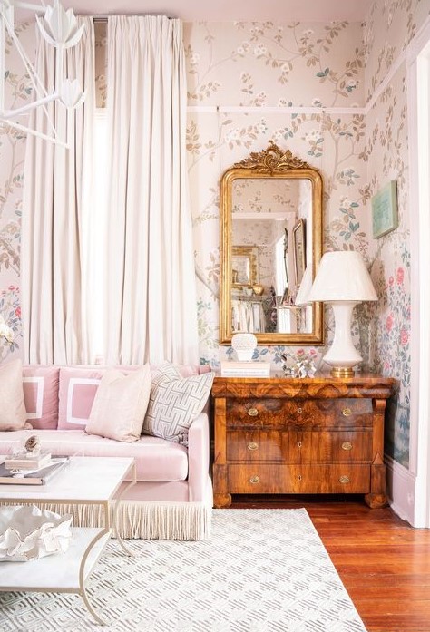 a catchy vintage living room with neutral floral wallpaper, a pink sofa with neutral pillows, a stained dresser, a mirror in a gold frame