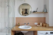 03 a mid-century modern bathroom with a bathtub, an open timber vanity with a washing machine, dusty pink tiles and a round mirror