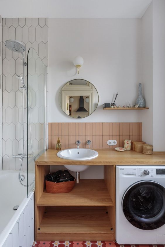a mid-century modern bathroom with a bathtub, an open timber vanity with a washing machine, dusty pink tiles and a round mirror