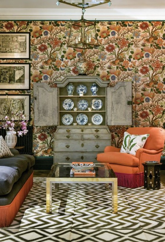 a maximalist cottagecore living room with bright floral wallpaper, a dark sofa and an orange chair, a black coffee table, a shabby chic buffet