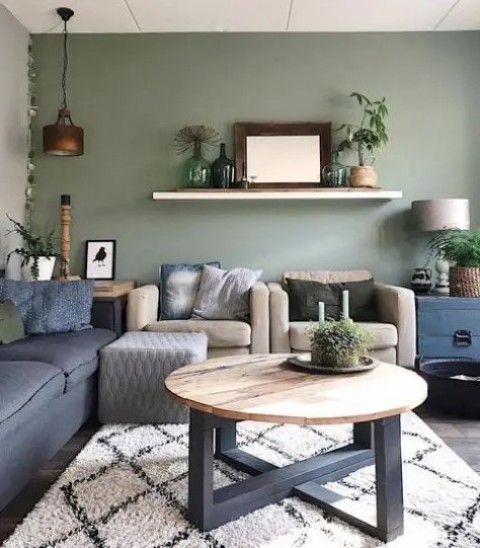 a modern boho living room with a sage green accent wall, a navy sofa and tan chairs, a round table and potted plants around