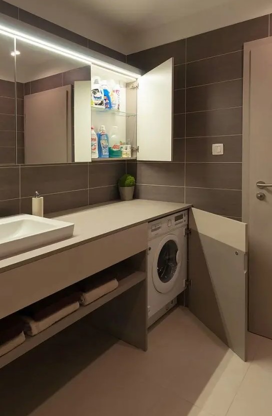 a modern taupe bathroom clad with large scale tiles, a sleek vanity that hides a washing machine, a large mirror cabinet