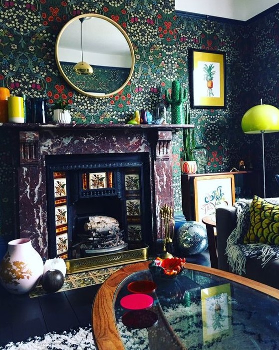 a moody living room with dark floral wallpaper, a non-working fireplace, a black sofa and bold pillows, a glass coffee table