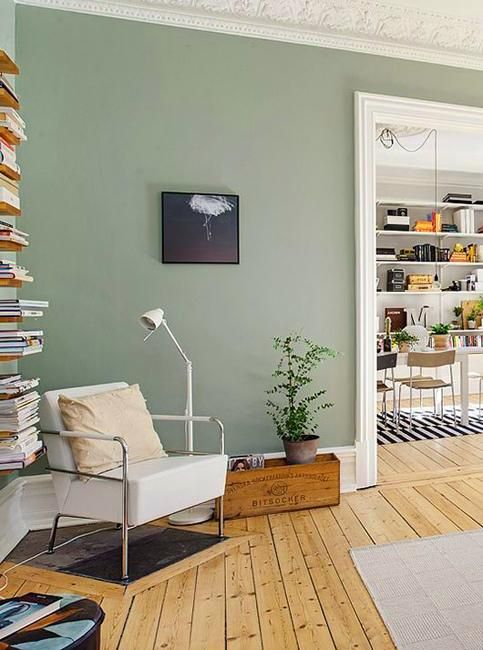 a Scandinavian living room with sage green walls, a neutral chair, an open shelf, a potted plant and some neutral textiles