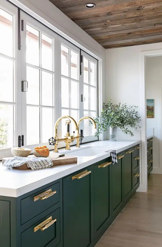 a hunter green modern kitchen with white stone countertops, gold handles and fixtures and a window instead of a backsplash