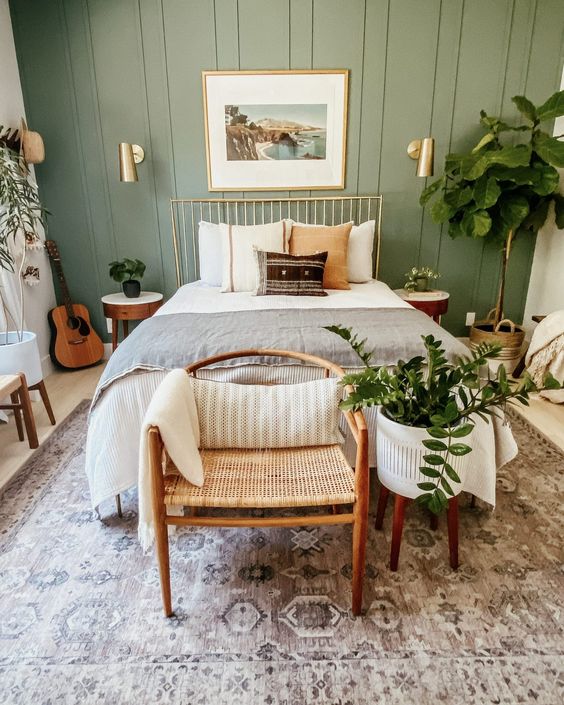 a modern bedroom with a sage green accent wall, a metal bed with neutral bedding, a woven chair, potted plants, gold touches for more elegance