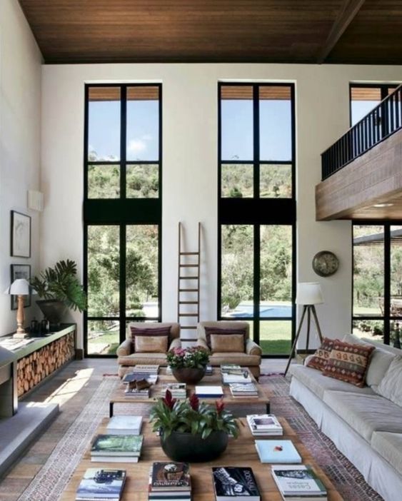 a neutral contemporary living room with double-height windows, neutral seating furniture, large coffee tables, a fireplace and a firewood storage unit