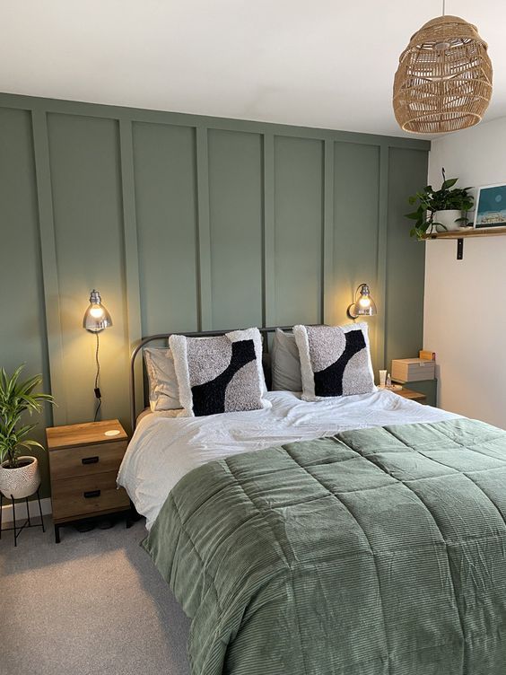 a modern bedroom with a sage green paneled wall, a metal bed with green and white bedding, stained nightstands, a woven pendant lamp