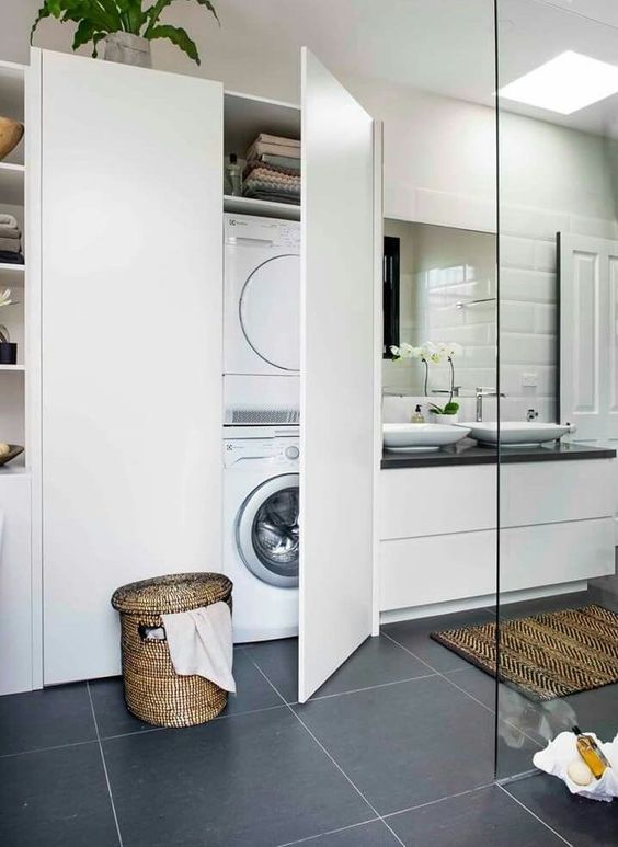 a modern bathroom clad with grey and white tiles, a white vanity with sinks, a large storage unit with a washing machine hidden