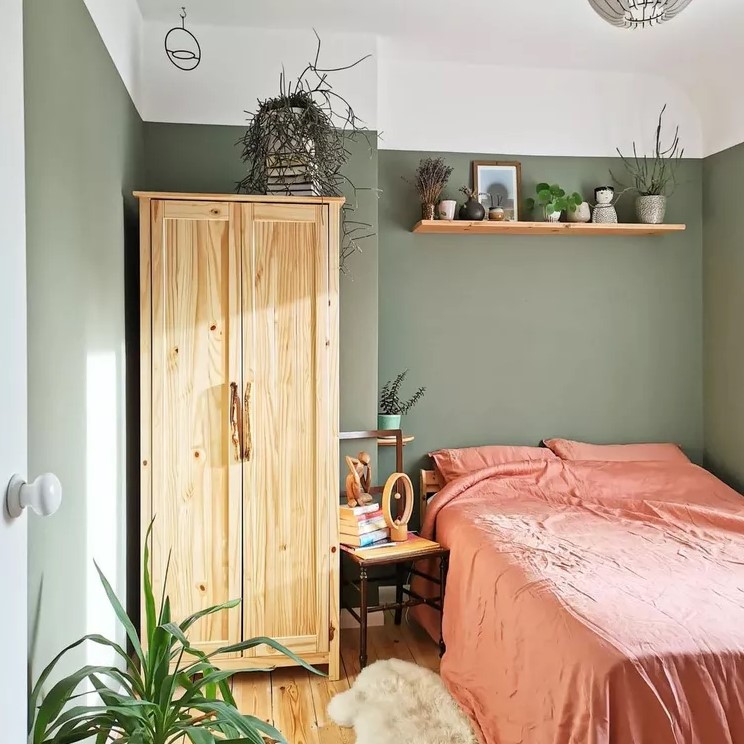 a small sage green bedroom with a stained wardrobe and a bed with coral bedding, open shelves, a chair and potted plants