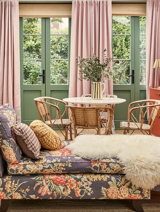 a muted color living room with green walls and doors, pink curtains, a floral daybed and a rattan dining nook