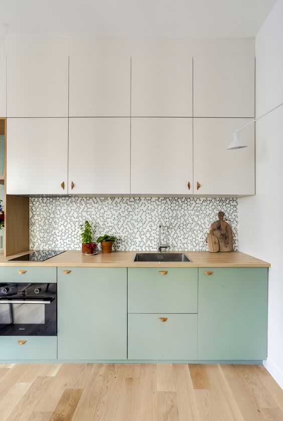 a two-tone kitchen with creamy and mint green cabinets, a printed tile backsplash, butcherblock countertops and built-in appliances