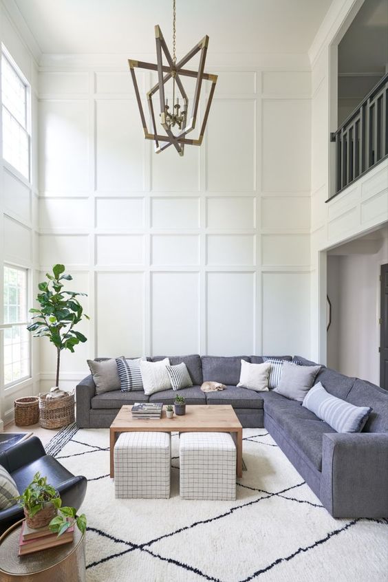 a neutral modern living room with trim on the walls, two rows of windows, a grey sofa, a table and some poufs and potted plants