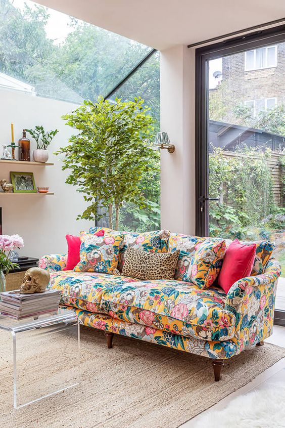 a neutral lviing room with glazed walls, a colorful floral sofa with bright pillows, a potted plant, a coffee table and shelves