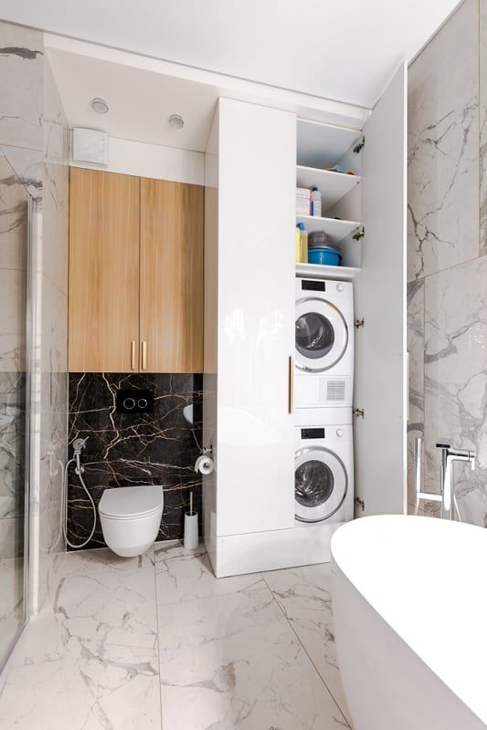 a small and chic bathroom clad with black and white marble tiles, a large storage unit with a washing machine and a dryer and a tub