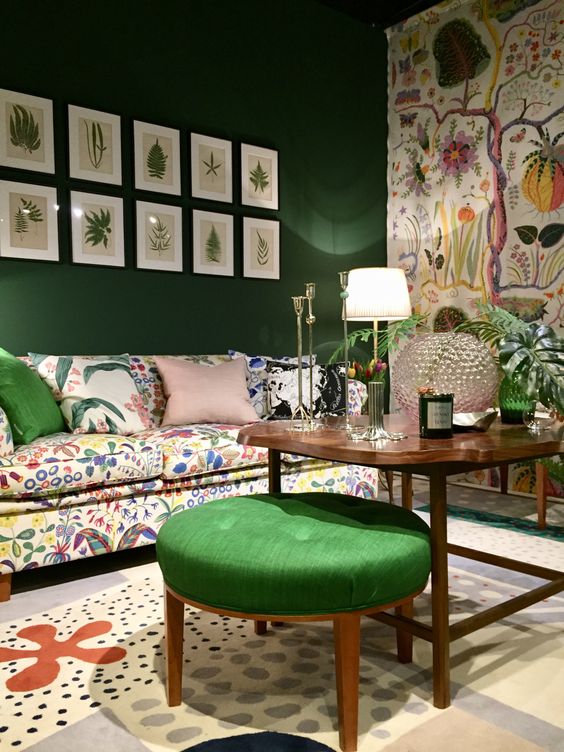 a whimsical living room with an emerald accent wall and a floral one, a crazy floral sofa and a rug, a gallery wall and a green pouf