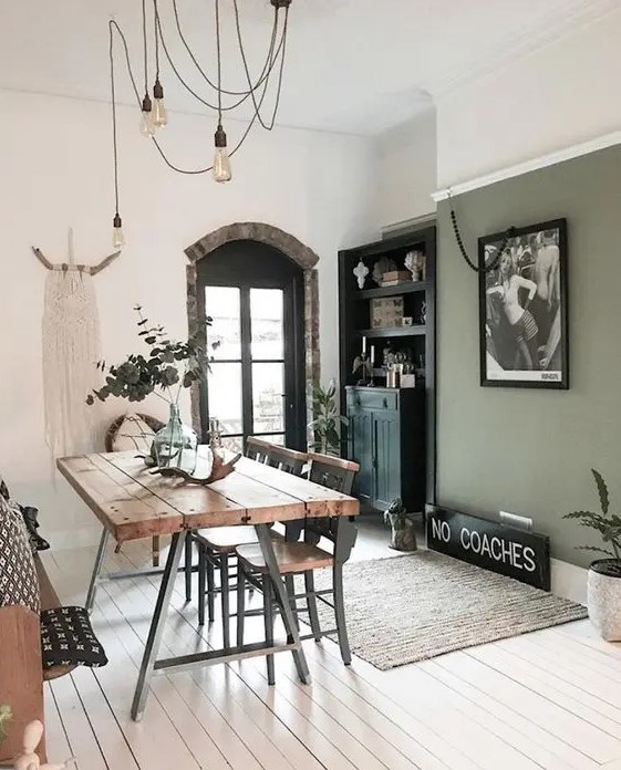 a boho dining room with a sage green accent wall, a black buffet, a trestle dining table, wooden chairs and bulbs plus macrame