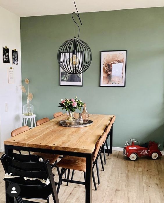 a modern dining room with a sage green accent wall, a stained table and leather chairs, a black pendant lamp and some art