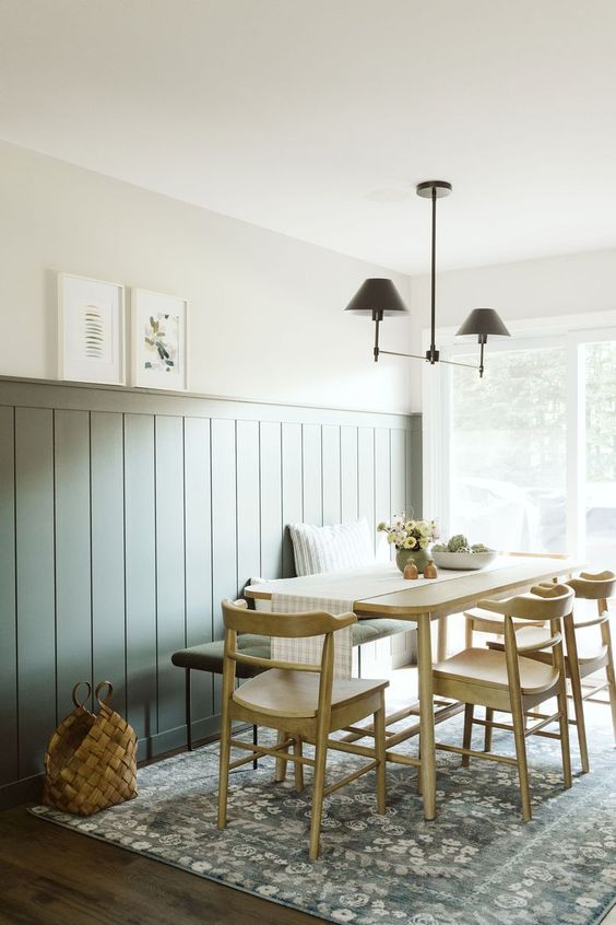 a modern dining room with sage green wall paneling, a stained table and chairs, an upholstered bench, a printed rug and a cool chandelier