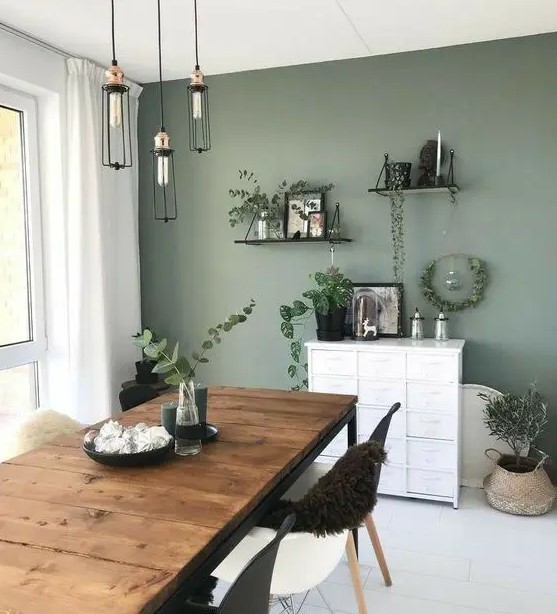 a Scandinavian dining room with a sage green accent wall, a rustic table, black and white chairs, pendant bulbs and open shelves