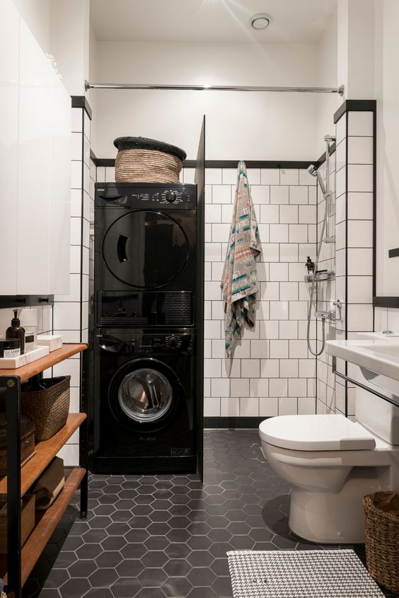 a Scandinavian bathroom with white square and black hex tiles, a wooden shelving unit, a shower and stacked washing machine and a dryer