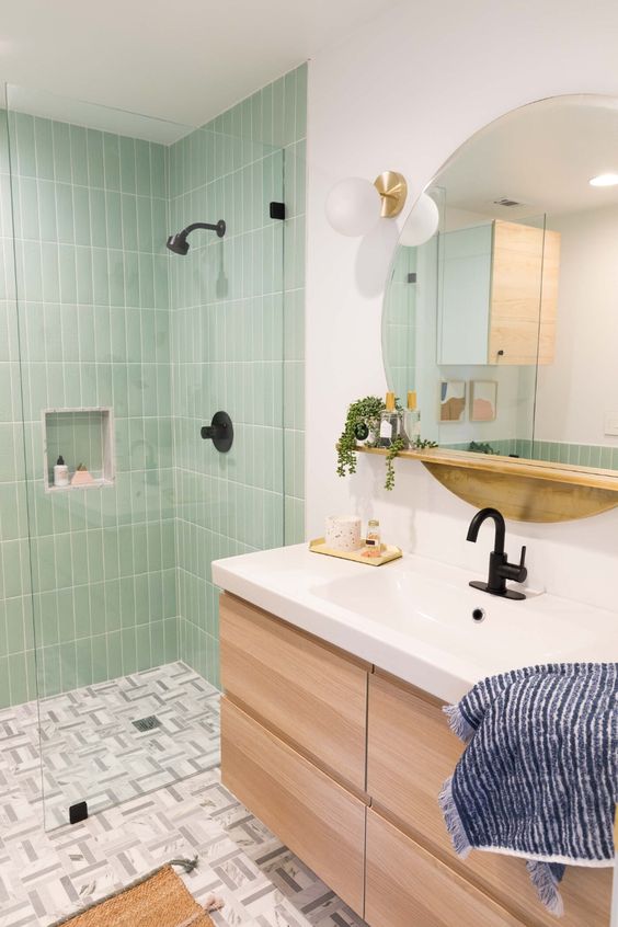 a farmhouse bathroom with mint green tiles in the shower space, a light-stained vanity, a round mirror and black fixtures