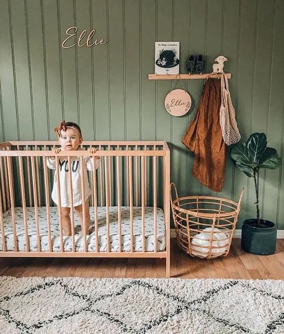 a cozy and simple nursery with a sage green beadboard accent wall, light stained furniture, a statement potted plant and some printed textiles