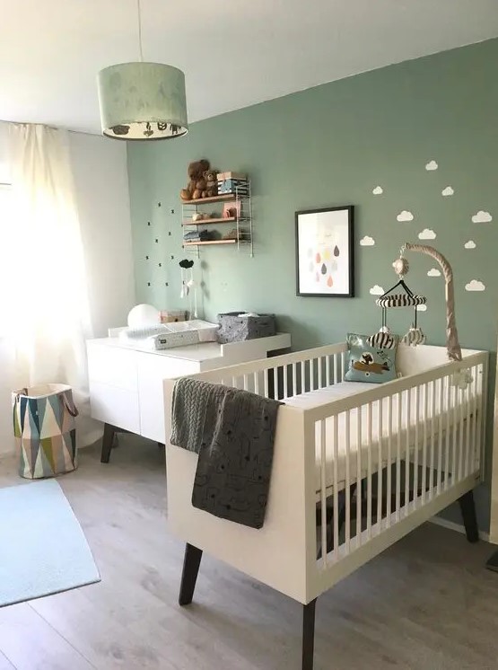 a cozy Nordic nursery with a sage green accent wall, white furniture, a pendant lamp and some shelves plus neutral bedding