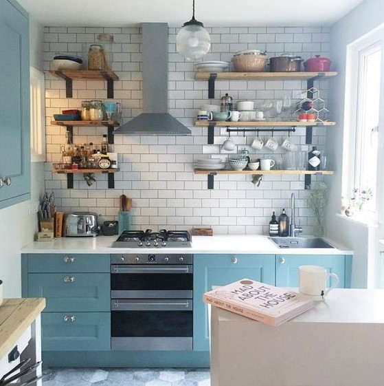 a small blue kitchen with a white tile backsplash and white countertops plus wooden shelves