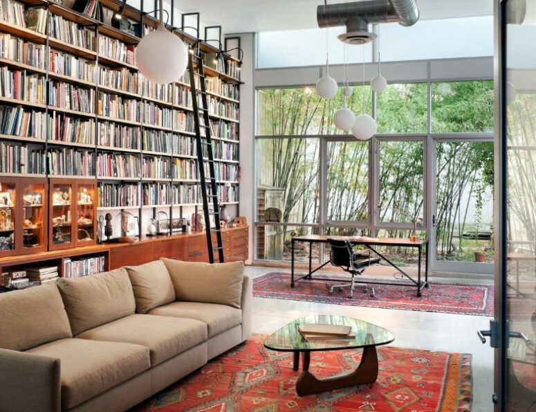 a home library with a double height ceiling and a whole wall taken by a bookcase, a desk by the glazed wall and a reading zone with a sofa