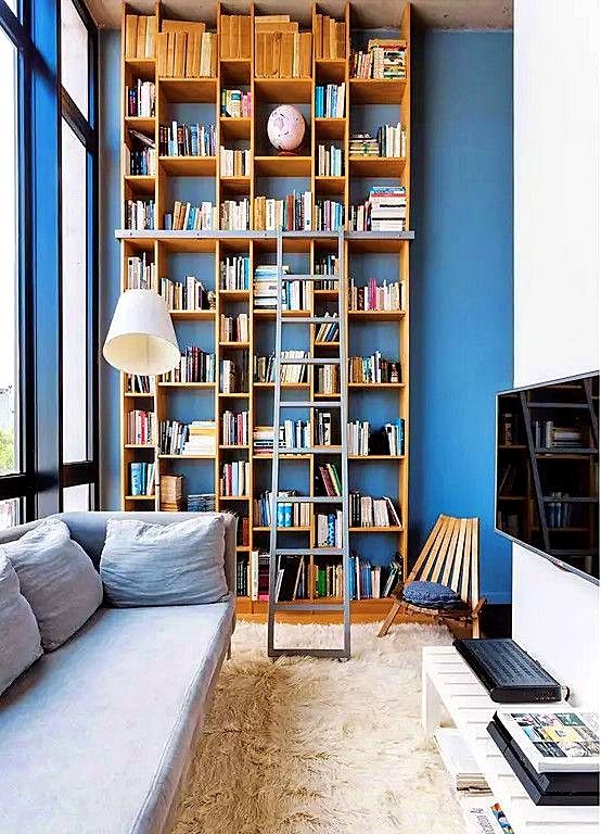 a narrow yet tall living room with a glazed wall, a blue accent wall, a grey sofa, a TV, a tall bookcase and a ladder plus a stained chair