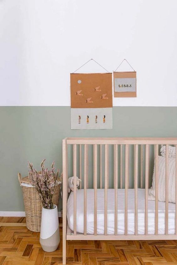 an airy and chic Scandinavian nursery with a color block sage green wall, a wooden crib, neutral bedding, a woven basket and a color block vase
