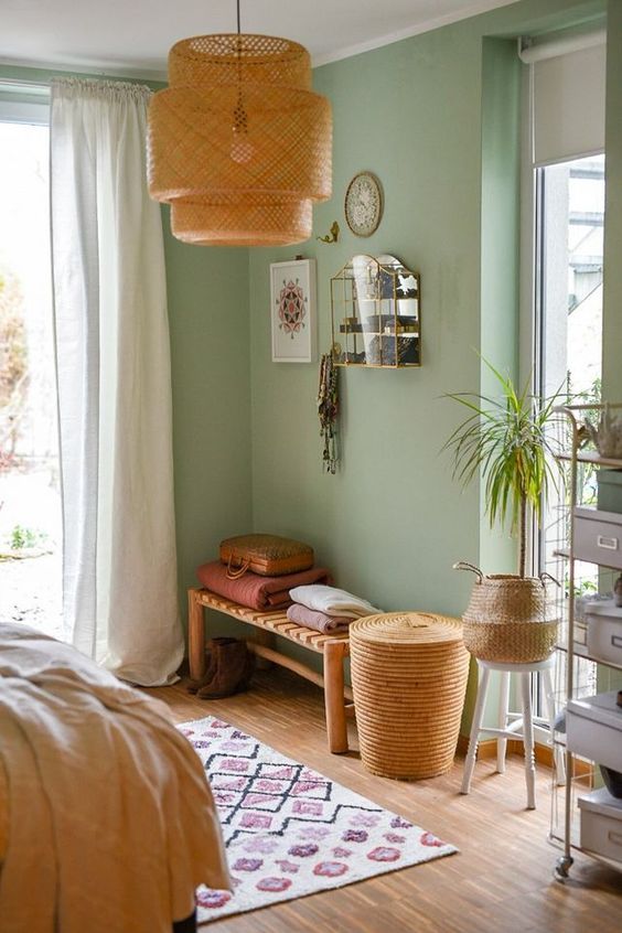 a mint green bedroom with neutral curtains, a woven bench and blankets, a shelving unit, baskets and a woven pendant lamp