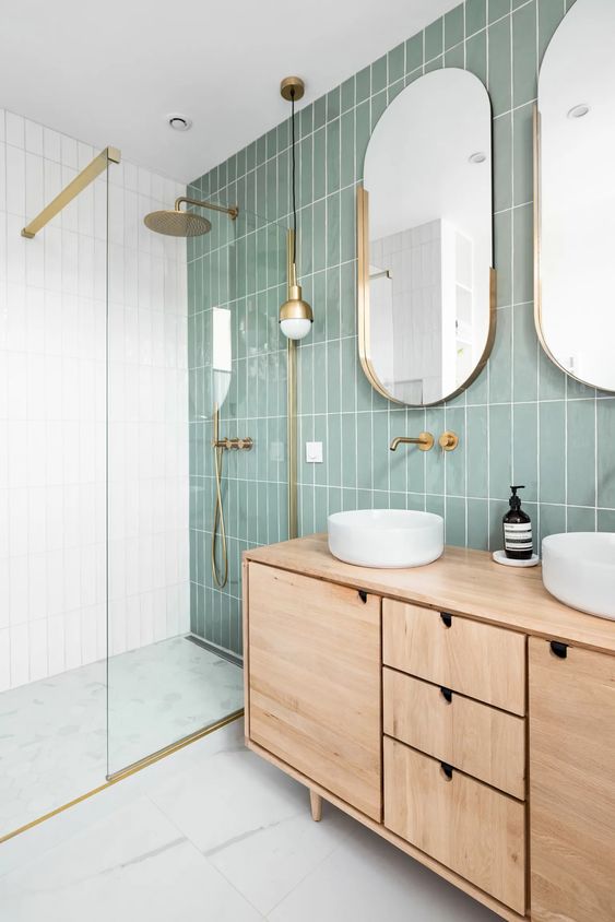 a modern bathroom with a sage green tile wall, a stained vanity, oval mirrors and round sinks, a shower space enclosed in glass