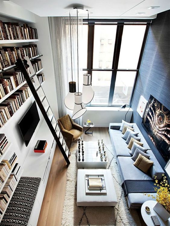 a small living room with a low sofa, a couple of coffee tables, a tall bookcase with a ladder, pendant lamps and neutral curtains