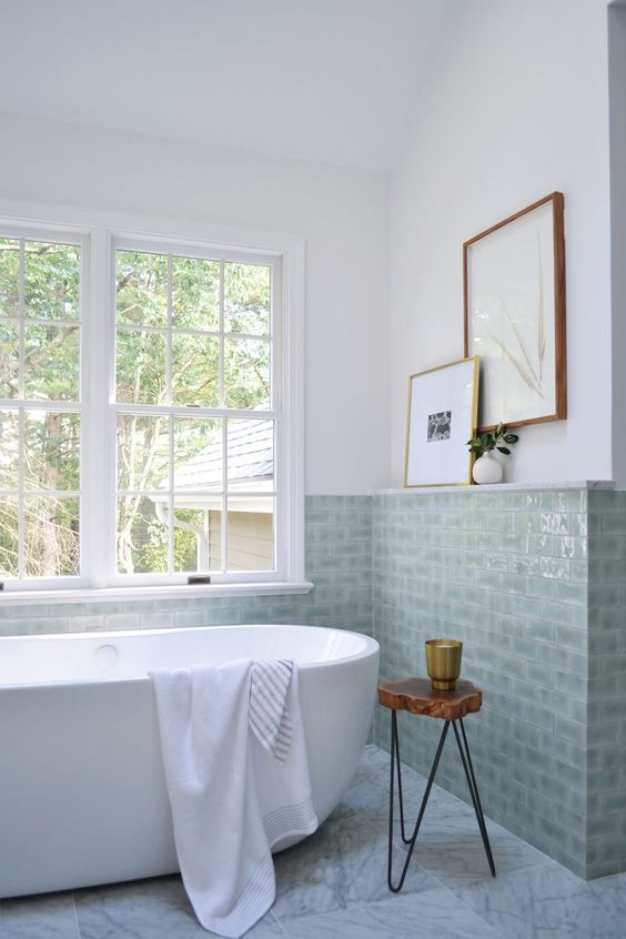 an airy bathroom in neutrals, with sage green tiles, an oval tub, a side table and artwork is a lovely and chic space