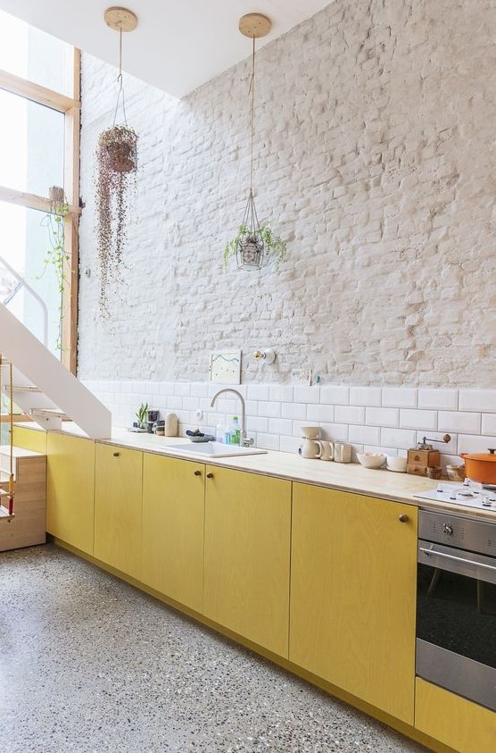 a bold contemporary kitchen with yellow plywood cabinets, neutral countertops, a white subway tile backsplash and a brick wall