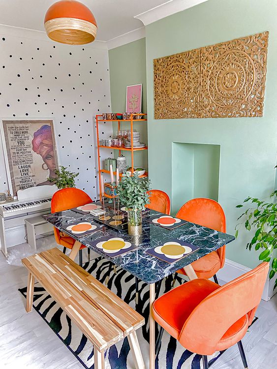 an eclectic boho dining room with a mint green accent wall, an orange shelving unit, orange chairs, a dark table and a bench