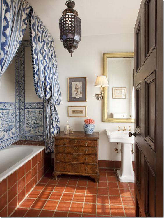 a Moroccan bathroom clad with terracotta tiles, with blue ones and blue curtains, a Moroccan pendant lamp and a dark-stained dresser