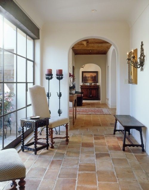 a Spanish style corridor with terracotta tiles, dark-stained and white upholstered furniture, tall candelabras and a glazed wall