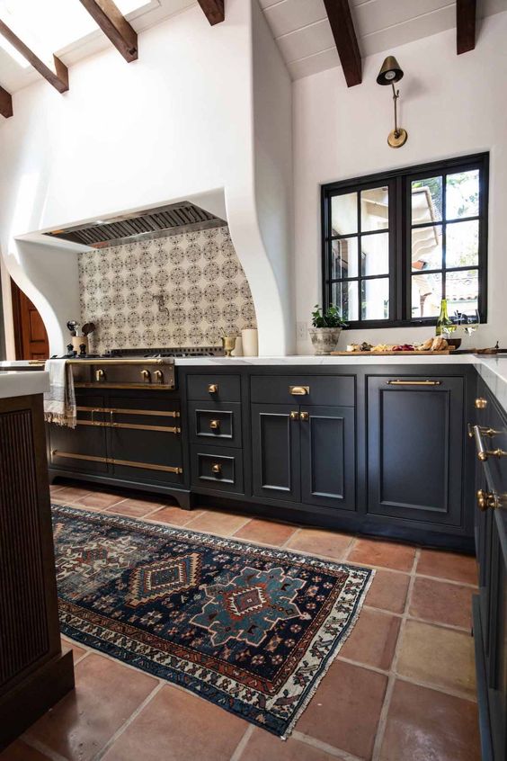 a Spanish style kitchen with a terracotta tile floor, navy cabinets, a large cooker and a built-in hood, dark-stained wooden beams