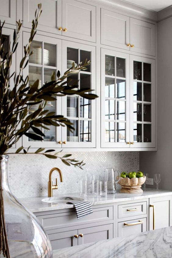 a beautiful dove grey kitchen with shaker and glass front cabinets, a tile backsplash and a white marble countertop plus gold handles