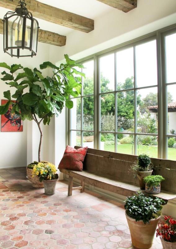 a beautiful entryway clad with hexagon terracotta tiles and a glazed wall, with a rough wood bench and beams, lots of potted plants