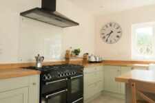 a beautiful sage green kitchen with butcherblock countertops, a black cooker and a hood, a glass backsplash is chic